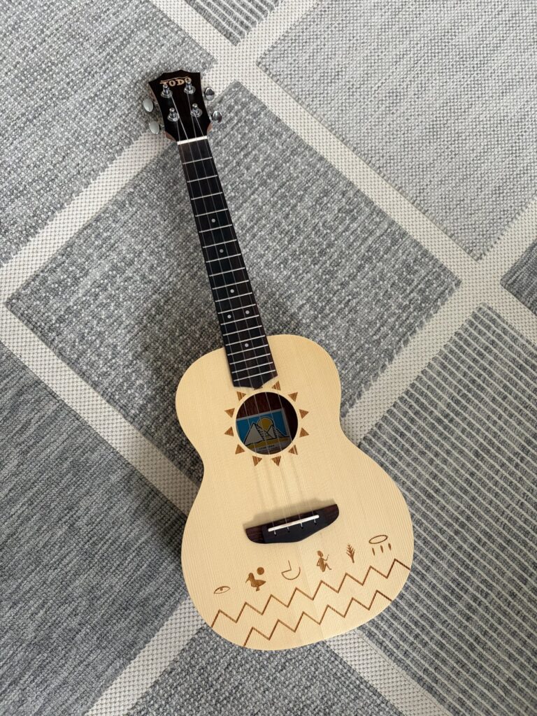 TODO The Nile Ukulele mit heller Decke, Sonnenmotiv und dunklem Griffbrett auf einem grauen Teppich