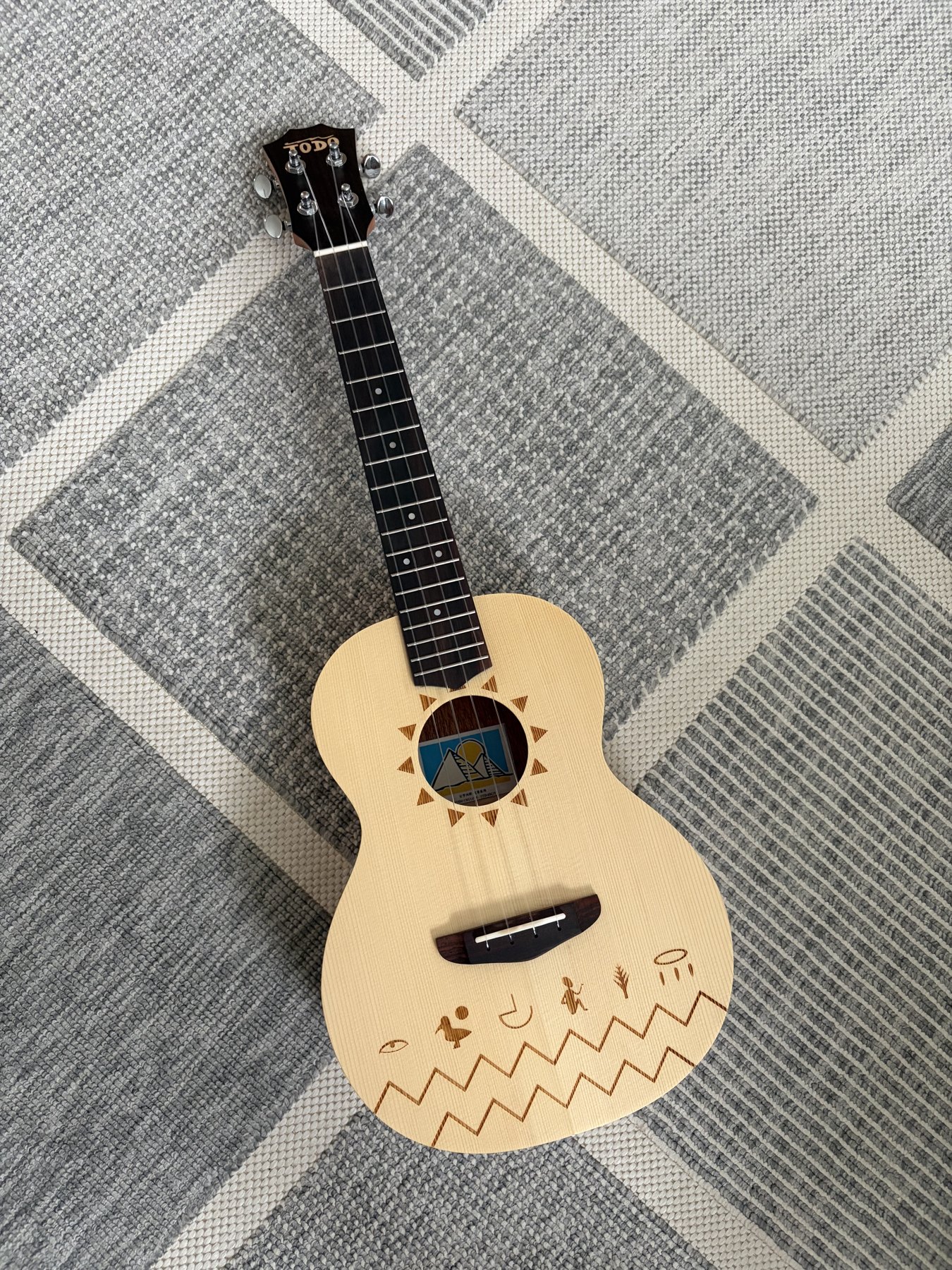 TODO Die Nile Ukulele mit heller Decke, Sonnenmotiv und dunklem Griffbrett auf einem grauen Teppich